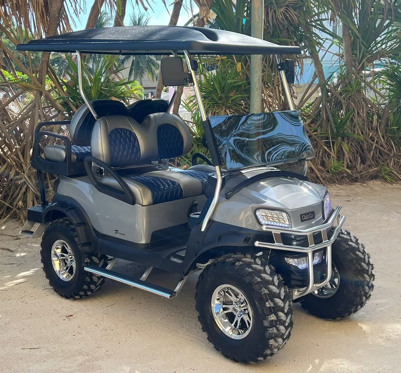 A silver lifted golf cart with a black roof and quilted seats parked on sand among tropical palms